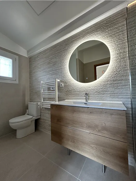 Rénovation de salle de bain à Sanary-sur-Mer avec meuble vasque effet bois, miroir rond rétroéclairé, revêtement mural texturé, sèche-serviettes blanc et WC au sol. Salle de bain contemporaine avec meuble bois et miroir lumineux