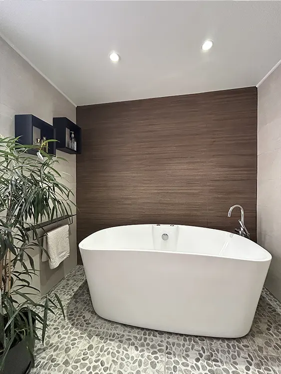 Baignoire îlot blanche installée devant un mur effet bois foncé, avec robinetterie chromée et sol imitation galets à Sanary-sur-Mer. Baignoire îlot et ambiance bois contemporaine